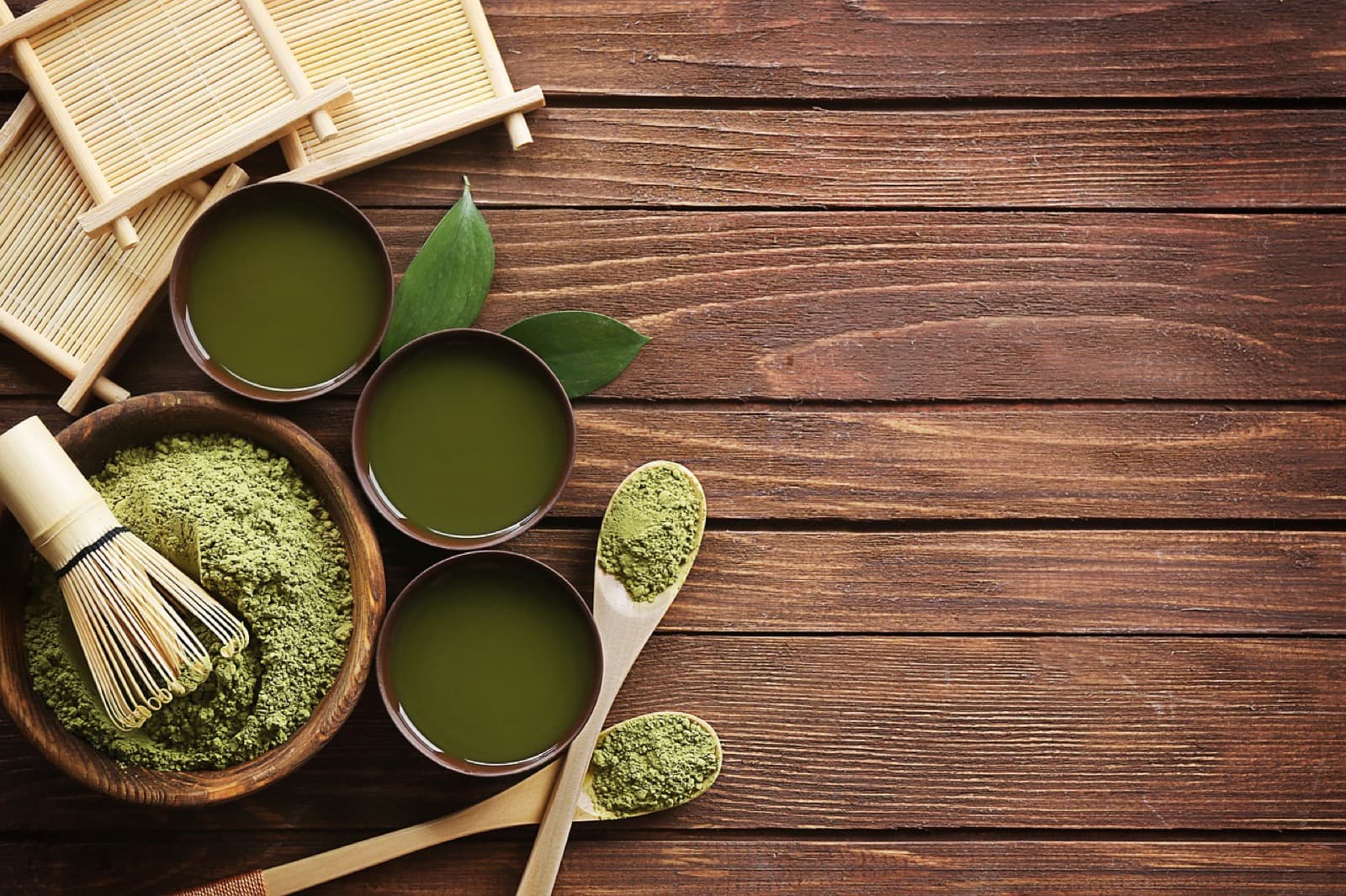 Matcha powder, bamboo whisk, and cups of whisked green tea on a wooden table
