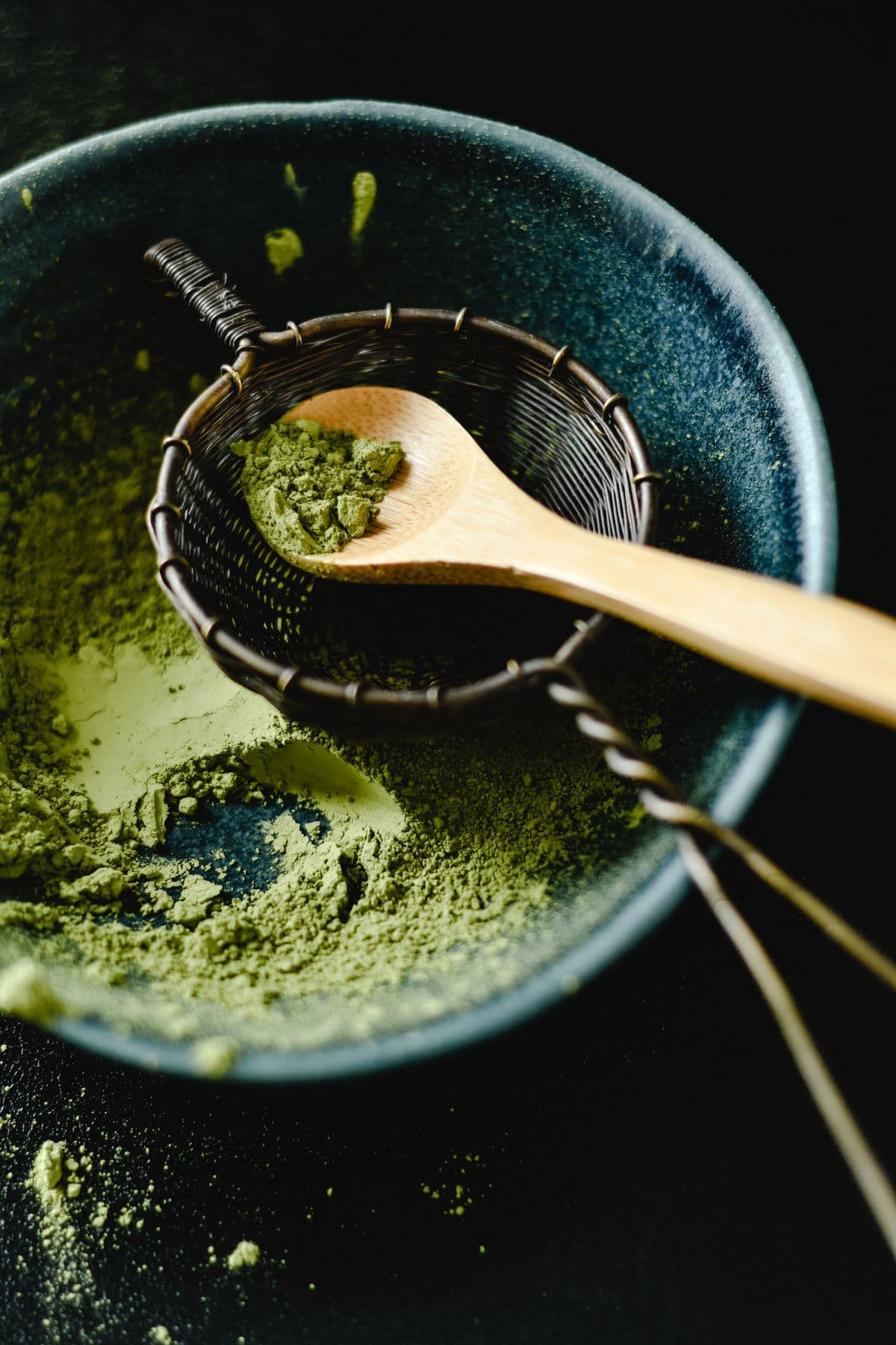 Matcha powder being sifted through a small stainless steel sieve into a bowl