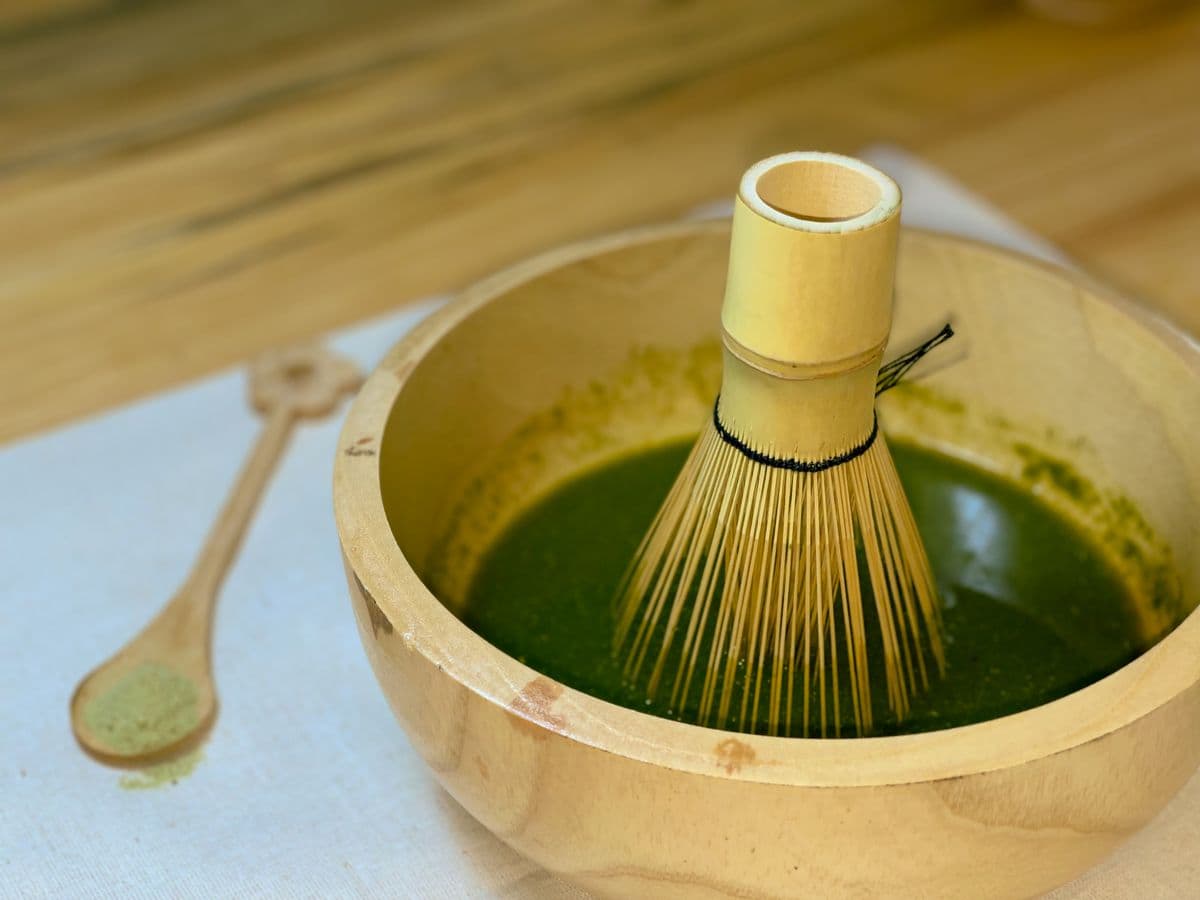 A bamboo whisk resting in a wooden bowl of freshly whisked matcha with a wooden scoop alongside