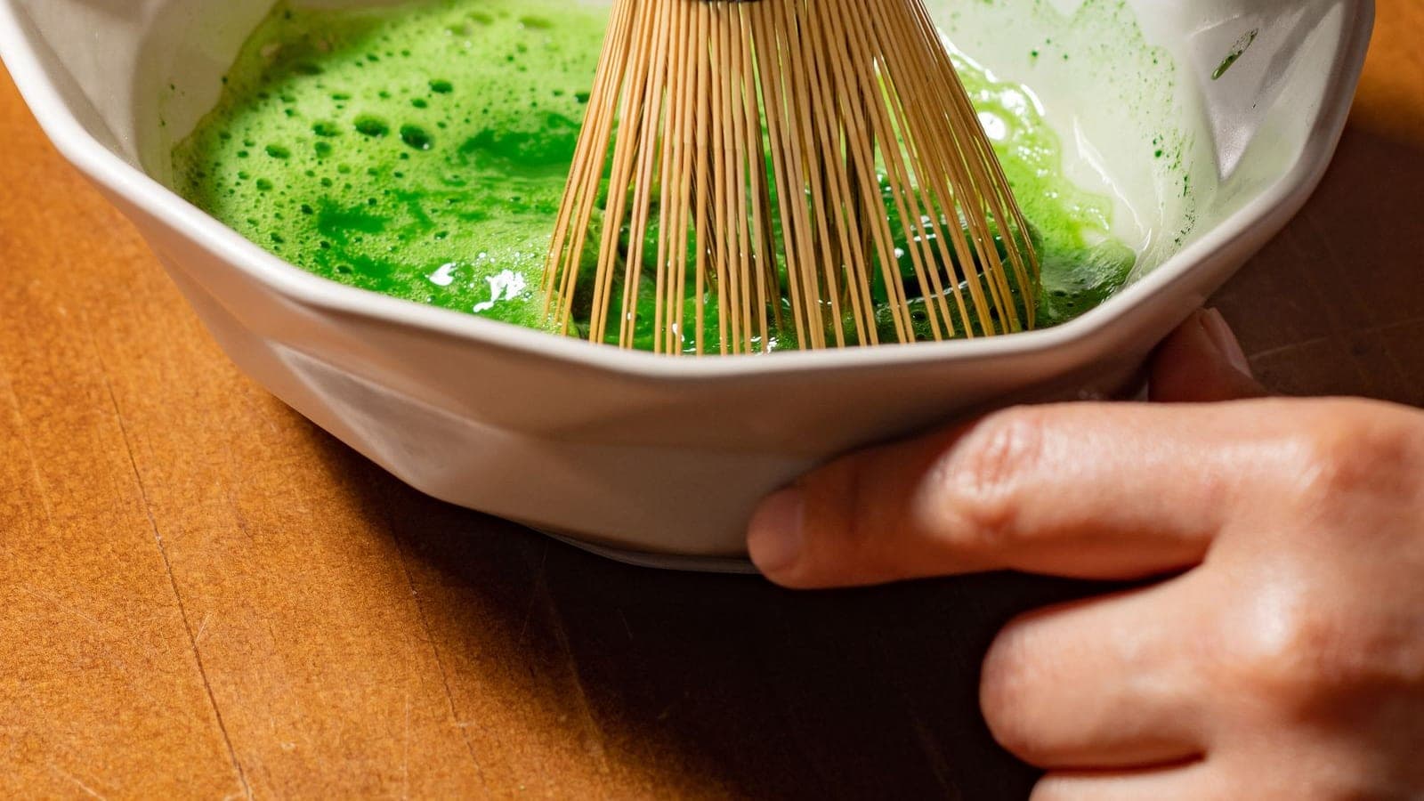 A hand steadying a white bowl while matcha is whisked to a bright green froth