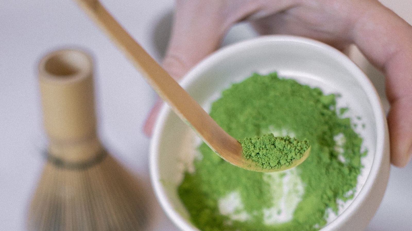 A bamboo scoop lifting vivid green matcha powder from a white bowl, with a whisk behind it